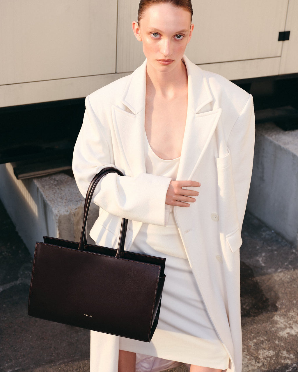 Poelle Atelier brown leather tote bag held by woman wearing white coat and dress outdoors