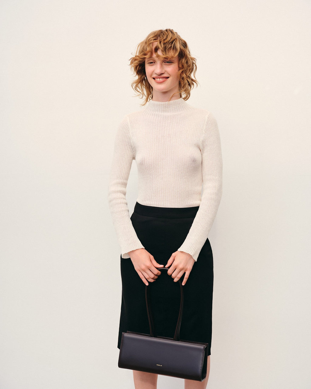 Woman holding a brown leather handbag by Poelle Atelier wearing a white sweater and black skirt