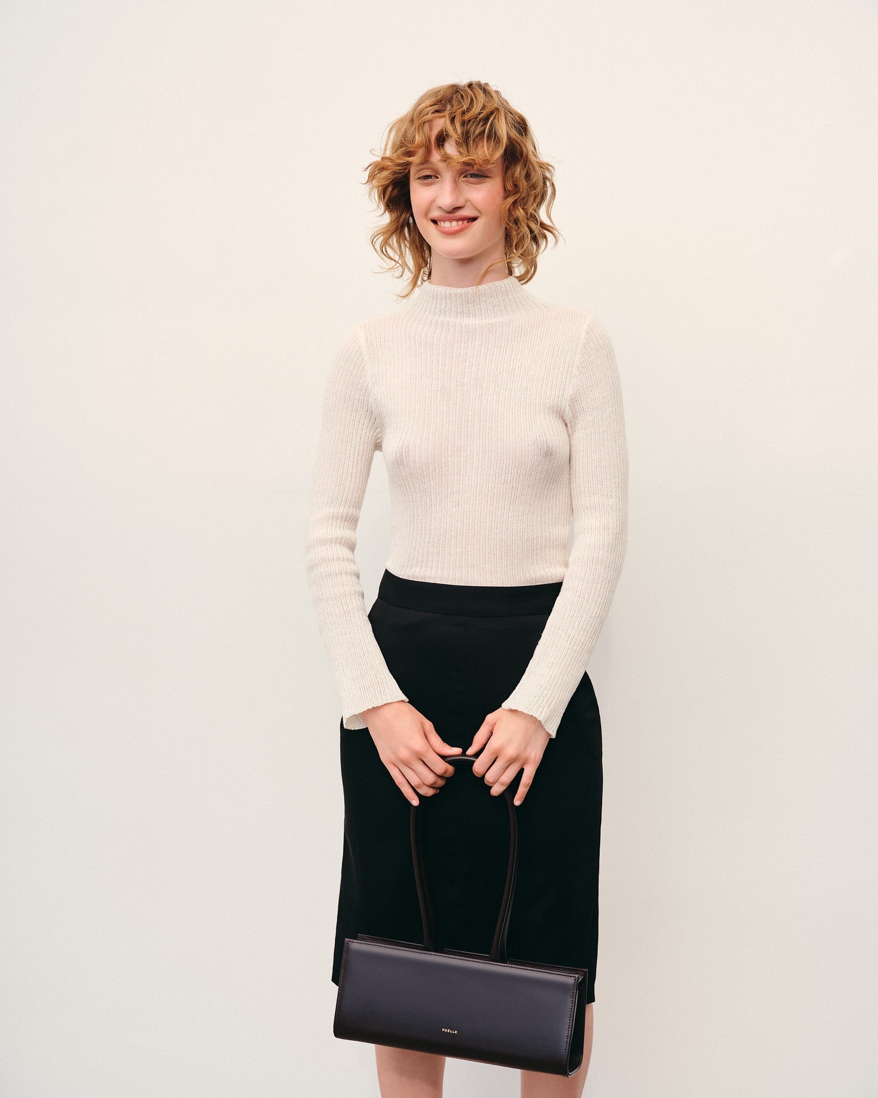 Woman holding a brown leather handbag by Poelle Atelier wearing a white sweater and black skirt