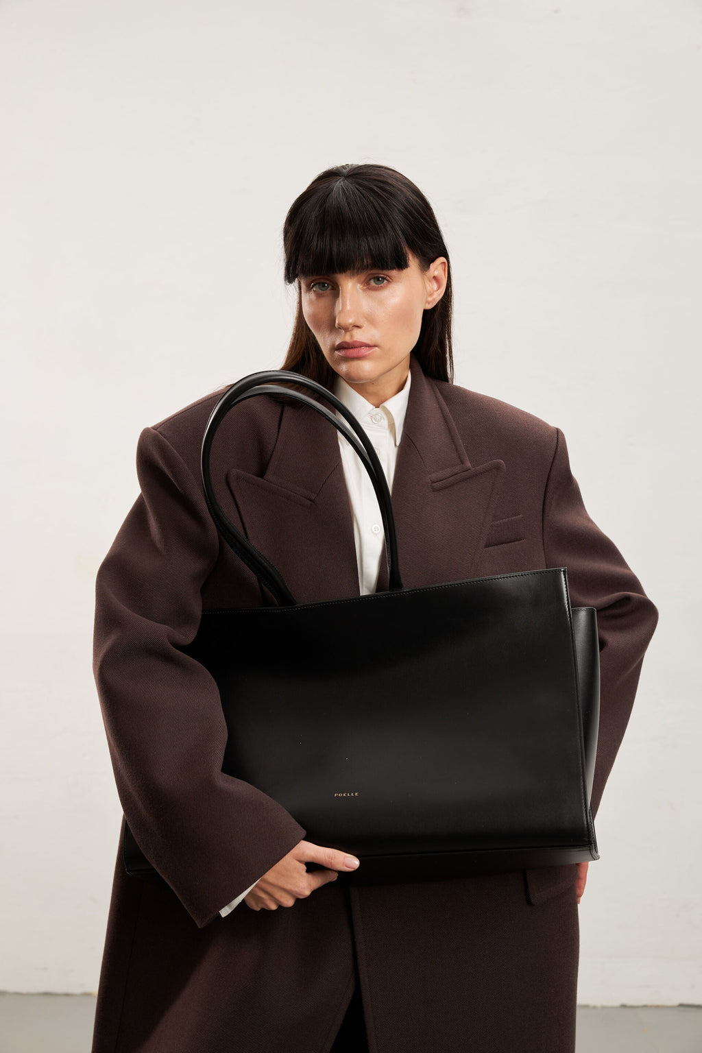 Luxury black leather tote by Poelle Atelier held by model in oversized brown coat