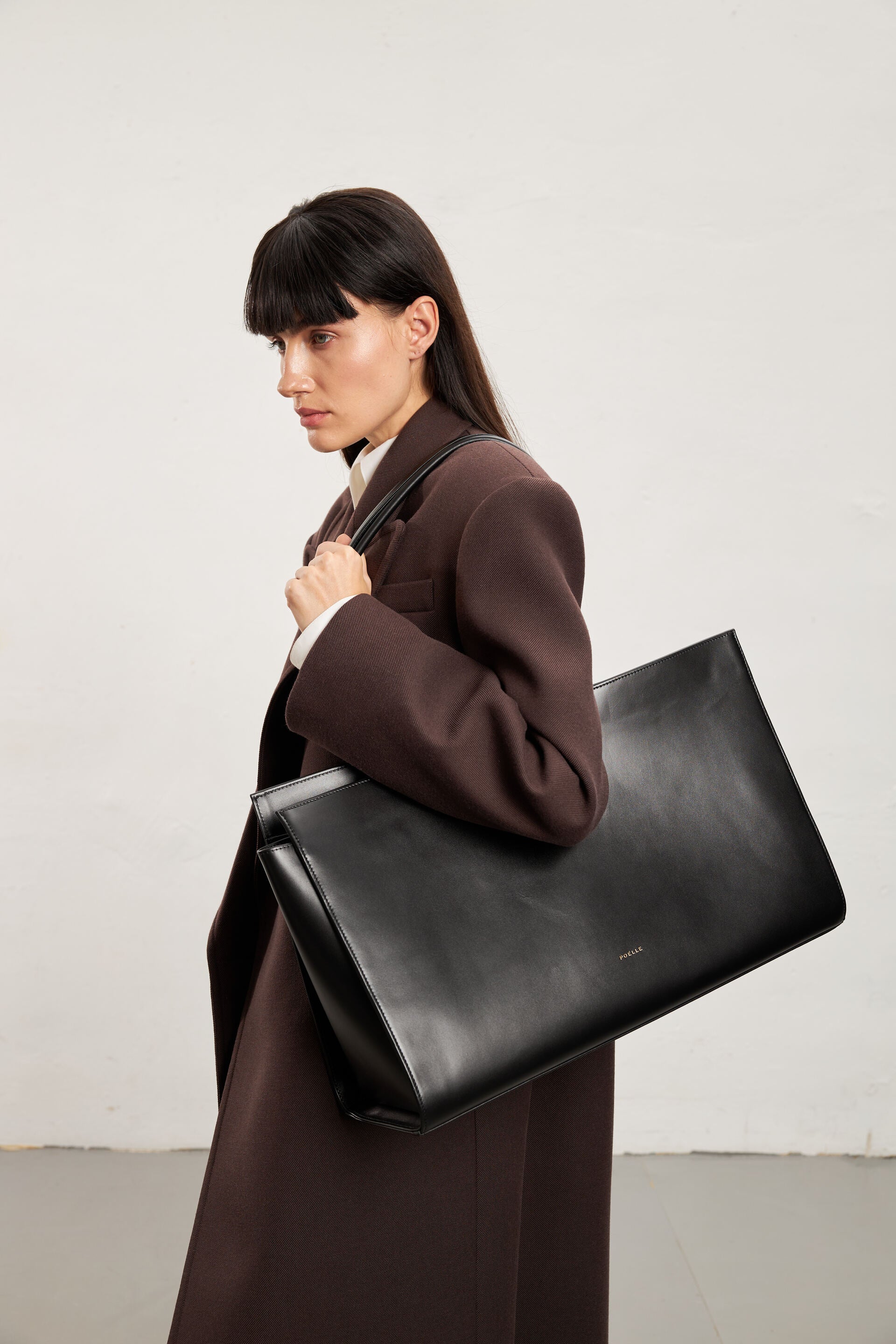 Woman carrying luxury black leather tote by Poelle Atelier in stylish brown coat